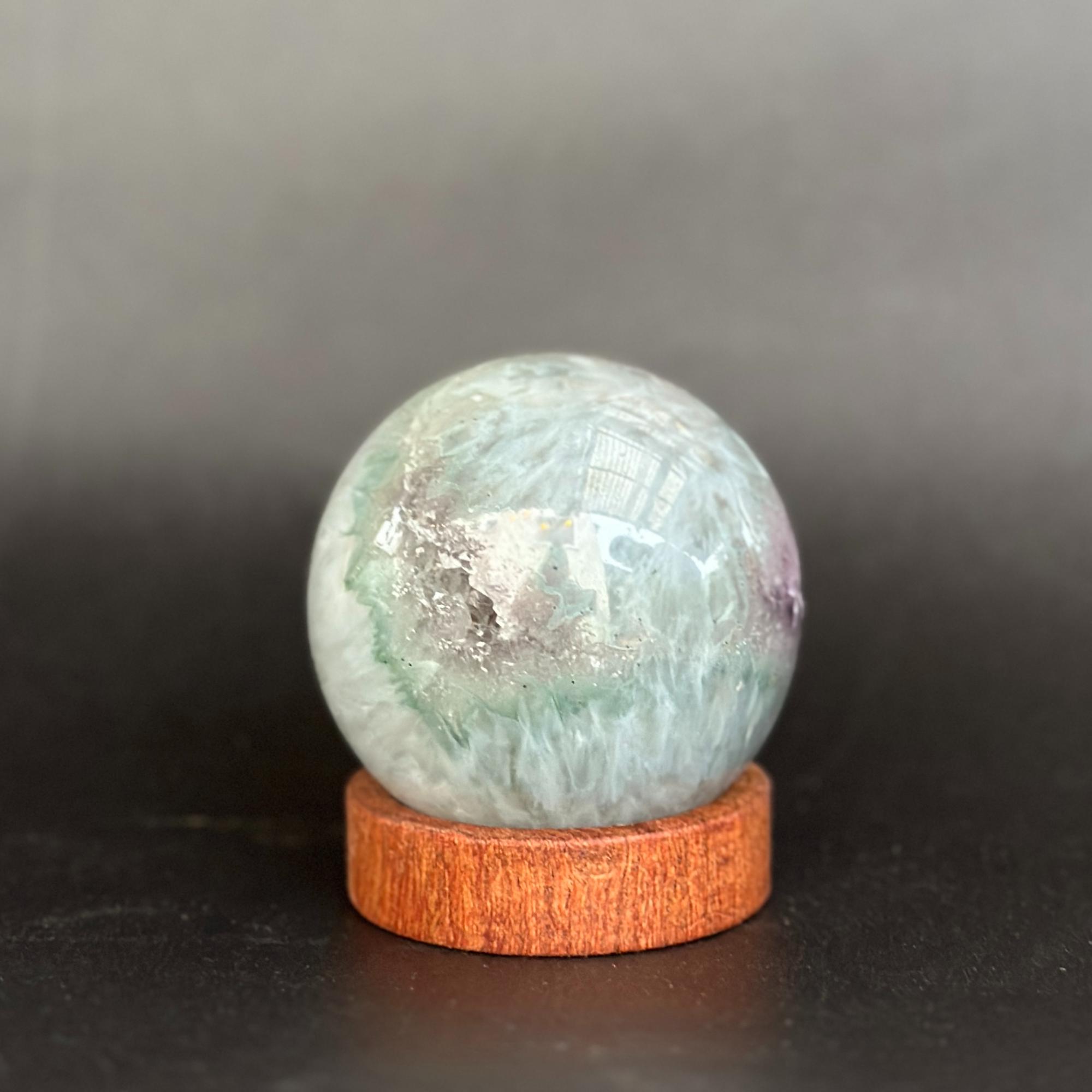 A polished round crystal ball with iridescent green, blue, white, and purple marbling, displayed on a small round wooden stand against a dark gray background.