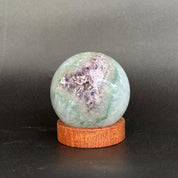 A polished crystal sphere with a geode-like interior showing purple and green hues, displayed on a round wooden stand against a dark gray background.