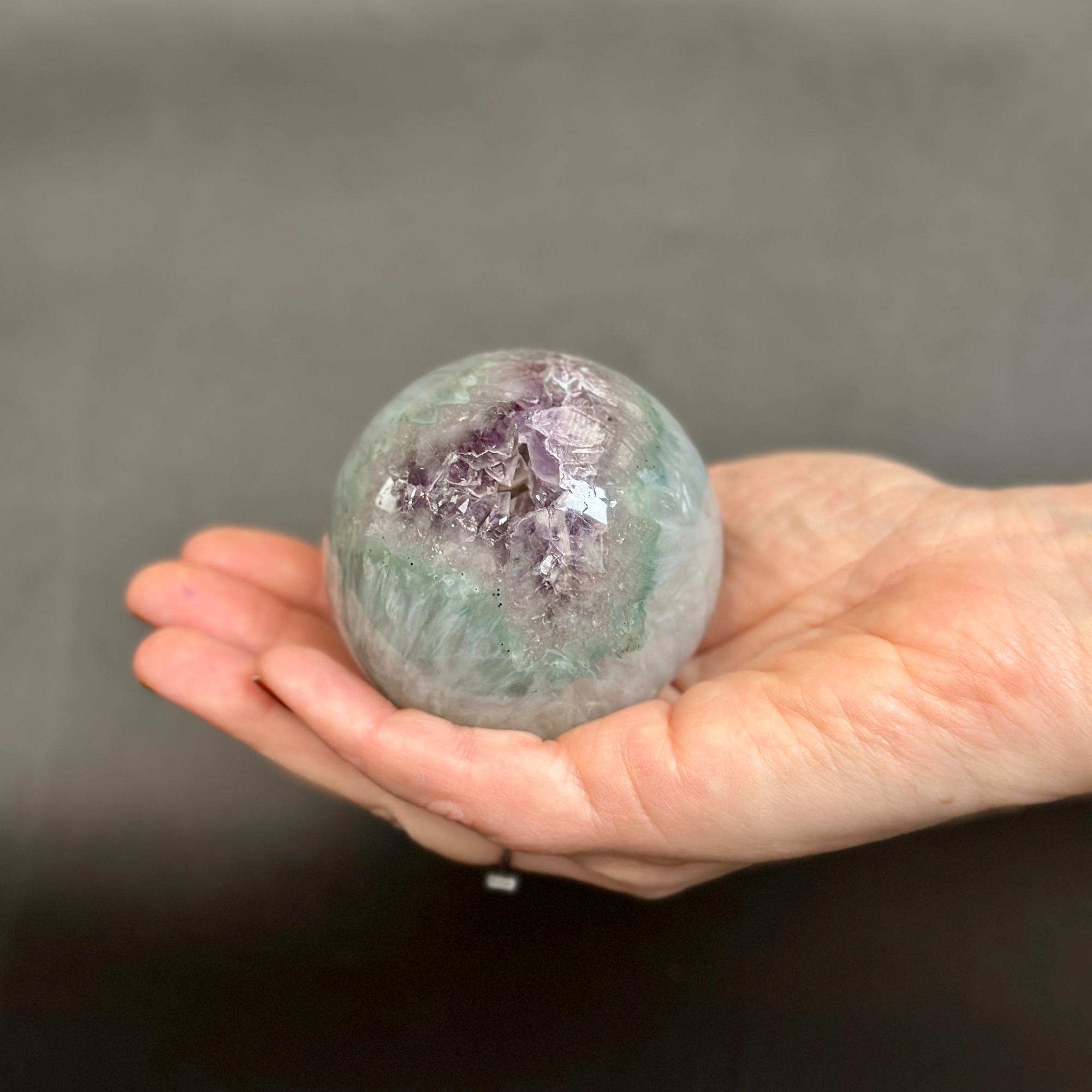 A hand holding a polished crystal sphere with a prominent purple amethyst cluster on one side and greenish-blue matrix on the other, set against a dark gray background.