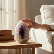 Person holding an amethyst geode on a wooden table with books and a plant in a bright room.