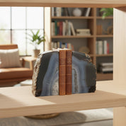Two decorative bookends shaped like sliced agate stones in dark gray, black, and blue-gray tones, with two brown leather-bound books standing in the center, displayed on a light wood shelf in a softly blurred home library setting.
