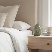 A styled bedroom scene with a neatly made bed in white linens and beige pillows beside a light wood nightstand holding a green jasper teardrop-shaped ceramic vase, a book, and a white mug.