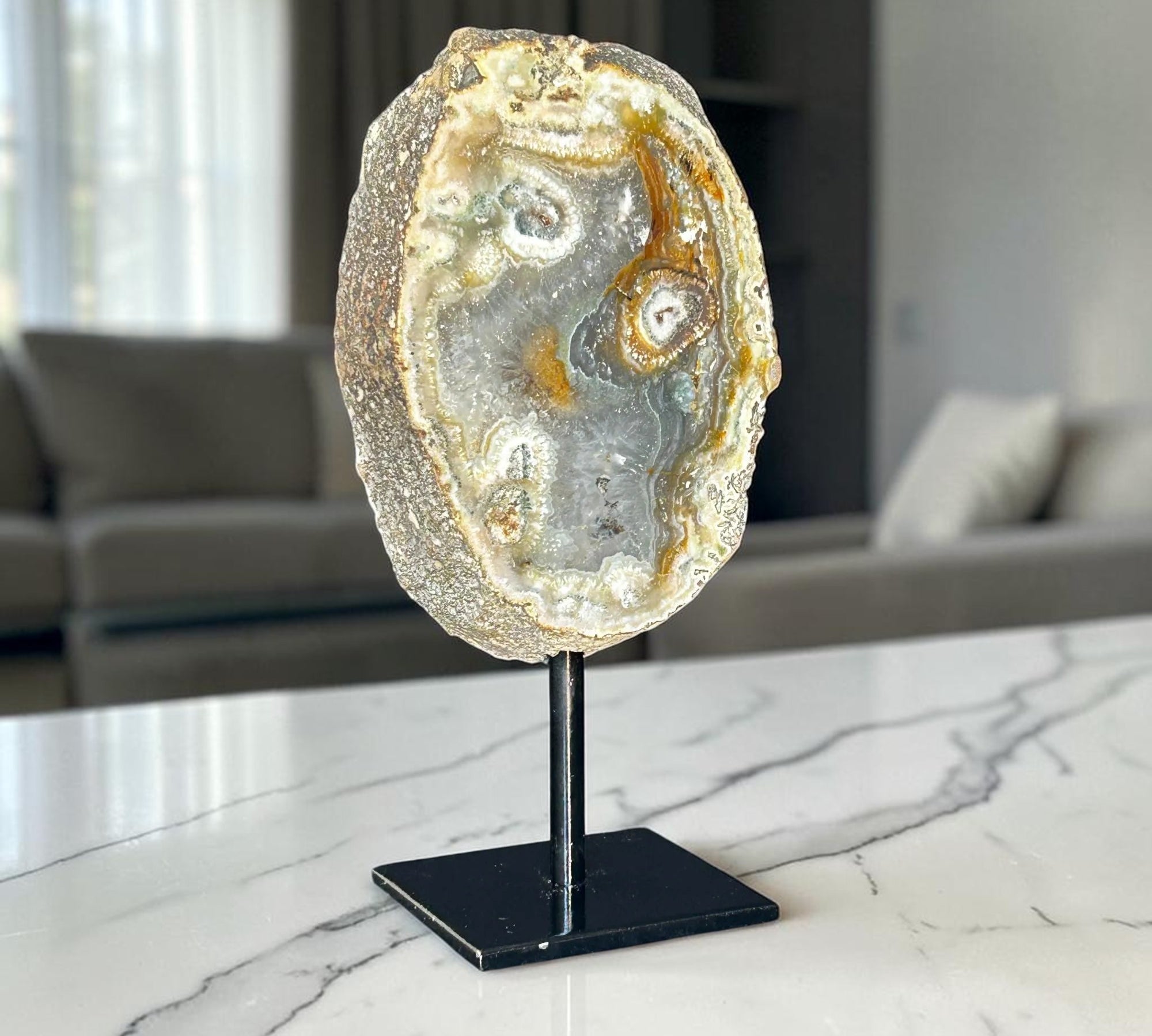 A polished agate and jasper geode slice on a black metal display stand, set on a white marble countertop with a blurred living room background.