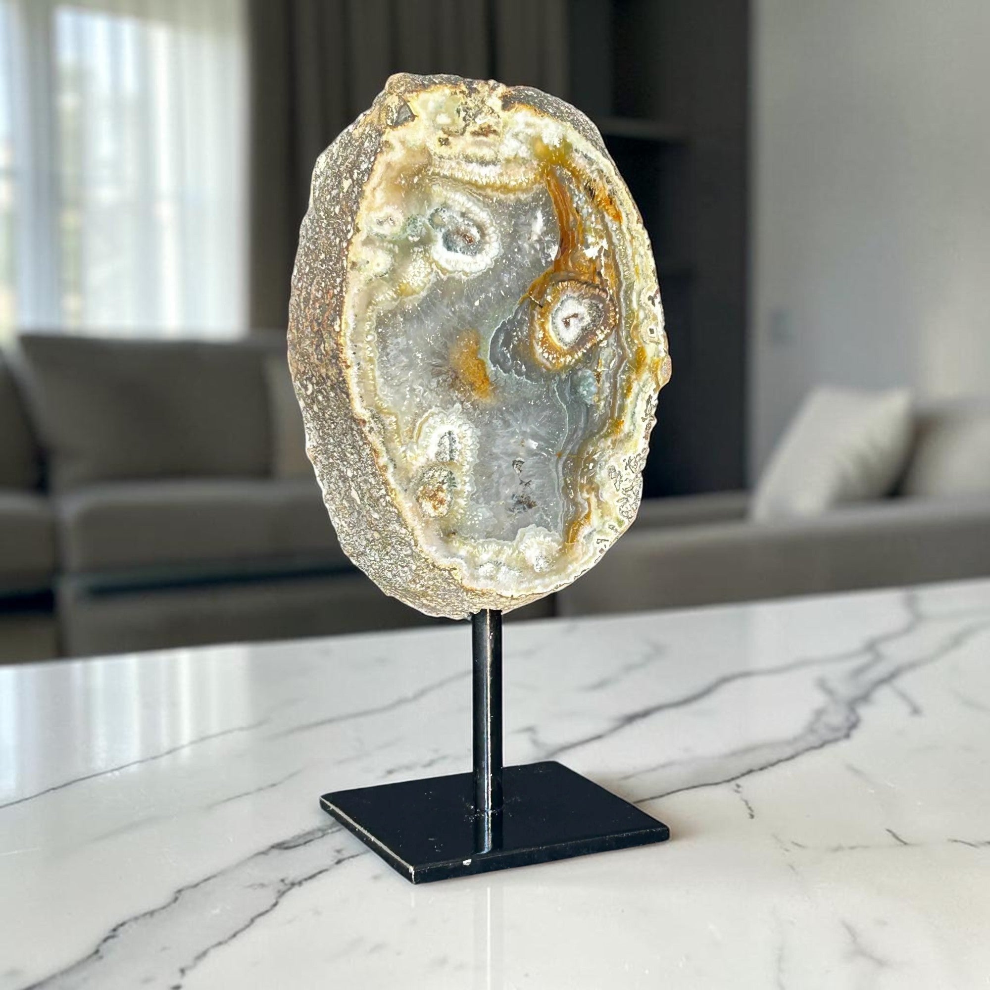 A polished agate and jasper geode slice on a black metal display stand, set on a white marble countertop with a blurred living room background.
