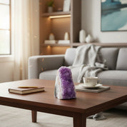 A purple amethyst crystal cluster sits on a dark wood coffee table in a modern living room setting, with a gray sofa, a cup of coffee, and a notebook in the background.