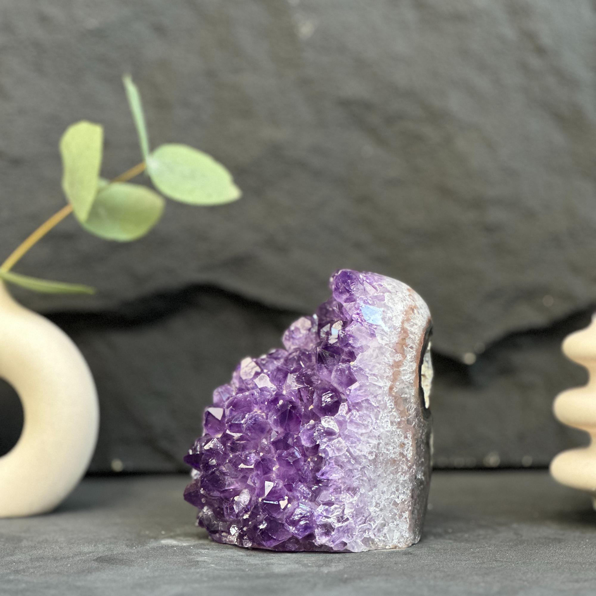 A natural amethyst crystal cluster geode specimen with deep purple crystals and a gray-brown matrix, displayed on a dark slate surface with decorative props.