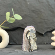 A small polished crystal or mineral specimen with purple, gray, and white marbling stands upright on a dark surface against a dark gray slate background, with partial decorative props visible at the left and right edges.