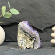 A raw amethyst geode specimen with a purple crystalline interior and a rough tan-brown outer matrix, displayed on a dark slate surface with blurred decorative props in the background.