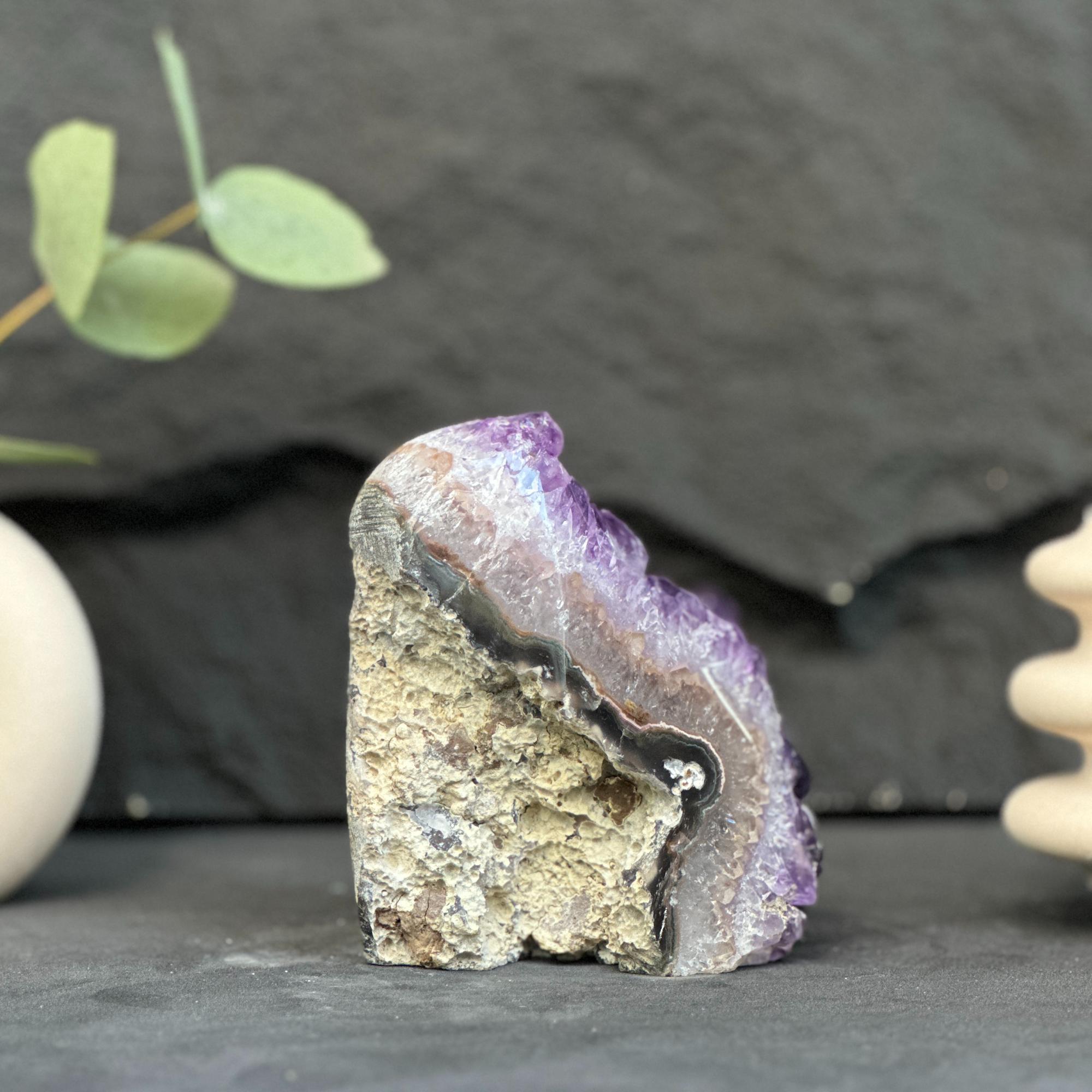 A raw amethyst geode specimen with a purple crystalline interior and a rough tan-brown outer matrix, displayed on a dark slate surface with blurred decorative props in the background.