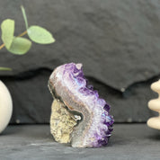 A natural amethyst crystal cluster geode specimen with purple and gray crystalline points on a tan matrix, displayed on a dark slate surface with decorative props in the background.