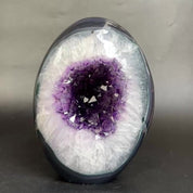 A polished geode-style decorative object with a dark outer shell and a bright white crystalline interior surrounding a deep purple crystal cavity, photographed on a dark neutral background.