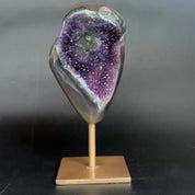 An amethyst and agate geode specimen on a wooden display stand, featuring a purple amethyst crystal cavity and green banded edges, set against a dark gray background.
