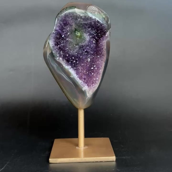 An amethyst and agate geode specimen on a wooden display stand, featuring a purple amethyst crystal cavity and green banded edges, set against a dark gray background.