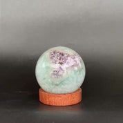 A round light blue-green crystal ball with white cloudy inclusions, displayed on a small round wooden base against a dark gray background.