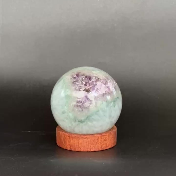 A round light blue-green crystal ball with white cloudy inclusions, displayed on a small round wooden base against a dark gray background.