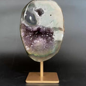 An amethyst and agate geode specimen on a wooden display stand, featuring a purple amethyst crystal cavity and green banded edges, set against a dark gray background.