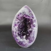 A polished amethyst geode specimen with a smooth egg-shaped outer layer and a deep, crystal-filled interior, photographed against a soft gray blurred background.