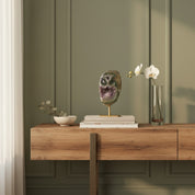A styled interior scene showing a wooden console table against a muted sage green paneled wall, with decorative objects including a geode sculpture, books, a small bowl, and a glass vase with white flowers.