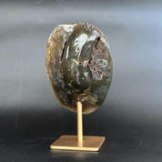 A polished natural stone specimen with a mottled green, gray, and brown pattern, mounted on a small gold-colored metal stand against a dark gray background.