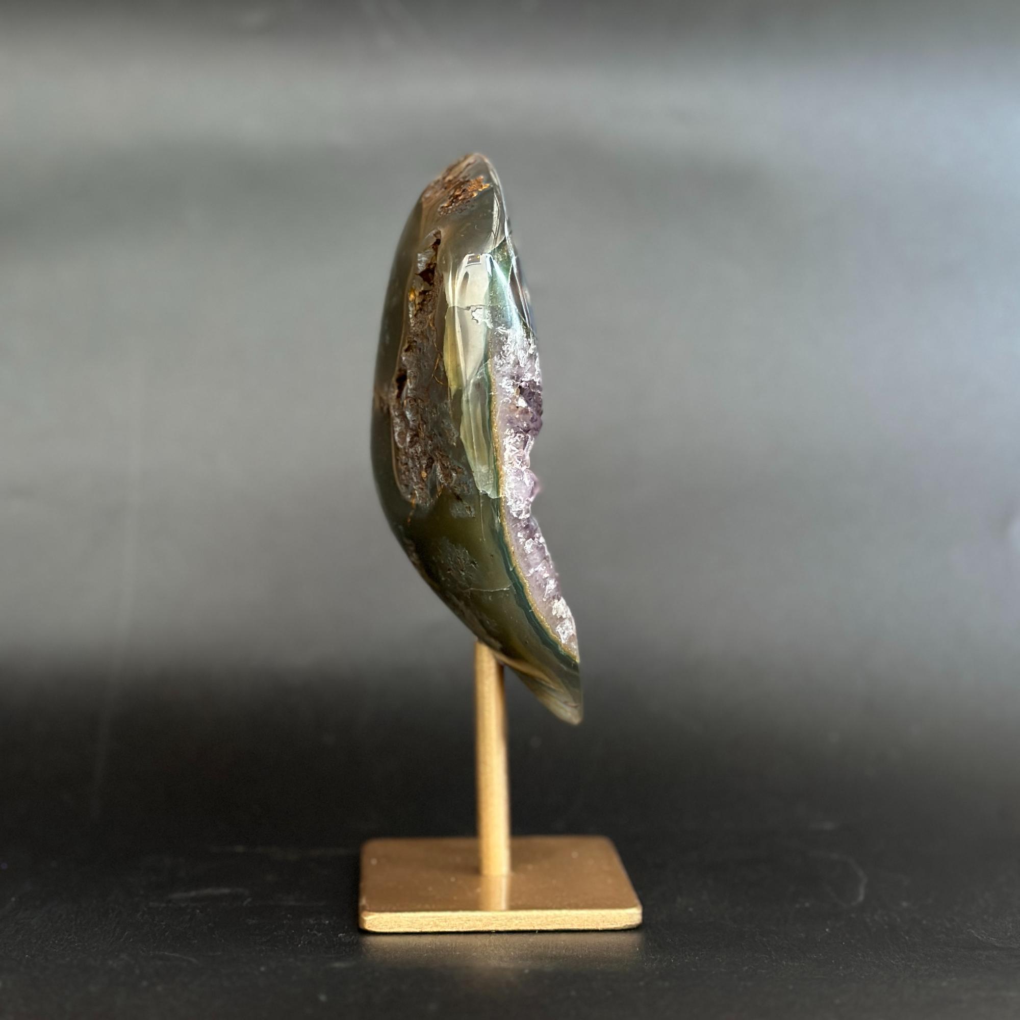 A polished natural stone or mineral specimen with an iridescent green and purple surface, mounted on a small wooden display stand against a dark gray background.