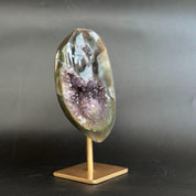 A polished natural amethyst geode slice with purple crystal formations, mounted on a small gold-colored metal stand against a dark gray background.