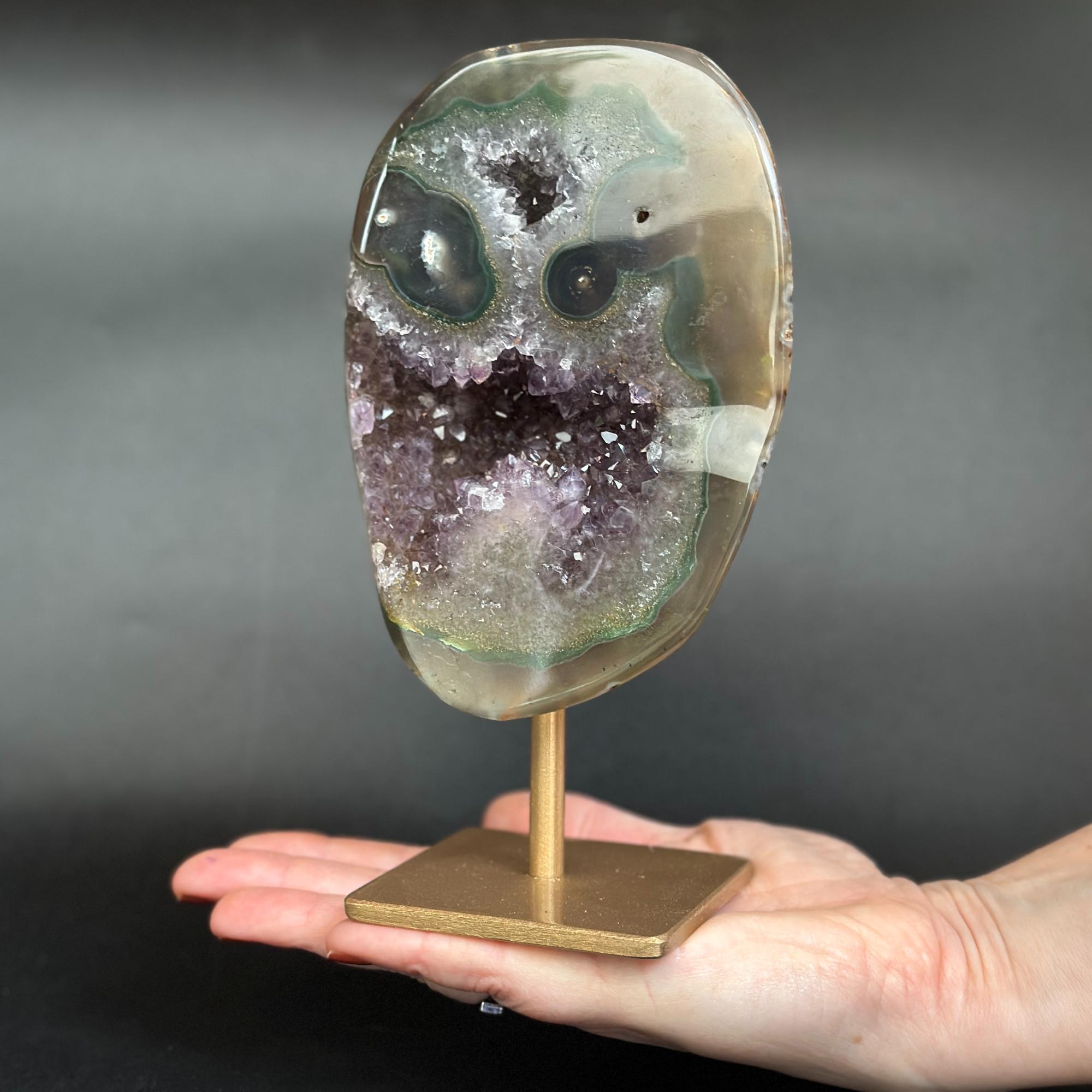 A polished agate geode crystal mount on a gold-colored metal stand, held in a person's hand against a dark gray background.