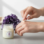 Person applying lotion to their hand next to a candle with a visible brand logo and amethyst